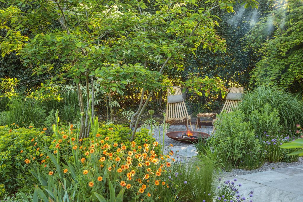 Sunken garden zone surrounded by planting and featuring two wooden garden chairs with a fire bowl in weathered steel.